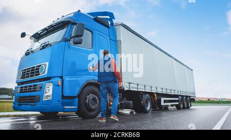 Le conducteur de camion traverse la route dans la zone rurale et entre dans son semi-camion bleu long Haul avec une remorque de cargaison attachée. Déménagement de la société de logistique Banque D'Images