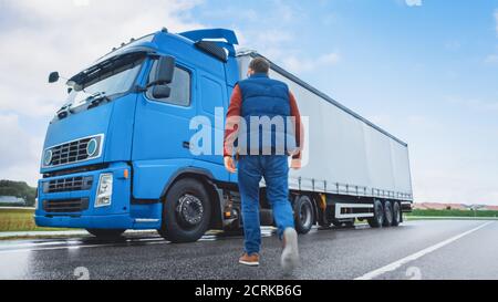 Le conducteur de camion traverse la route dans la zone rurale et entre dans son semi-camion bleu long Haul avec une remorque de cargaison attachée. Déménagement de la société de logistique Banque D'Images