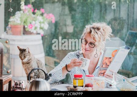 Blonde jeune femme mûre avec des verres en pyjama à la maison à l'heure du petit déjeuner, la lecture d'un magazine et d'avoir une tasse de café et chat. Banque D'Images