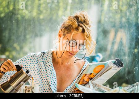 Blonde jeune femme mûre avec des verres en pyjama à la maison à l'heure du petit déjeuner, la lecture d'un magazine et la préparation d'une tasse de café. Banque D'Images