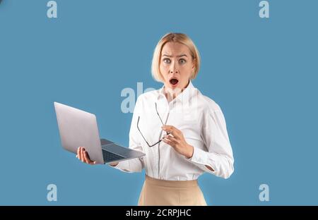 Portrait d'une femme d'affaires choquée tenant un ordinateur portable et regardant l'appareil photo sur fond de studio bleu Banque D'Images