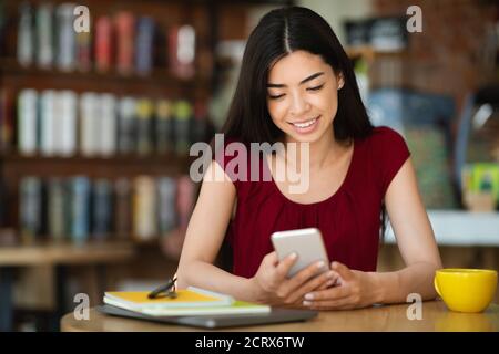 Une femme asiatique souriante envoie des SMS sur un smartphone tout en prenant une pause café Au café Banque D'Images