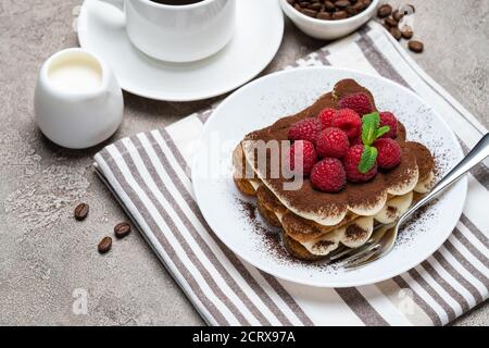 Portion de dessert tiramisu classique avec framboises et une tasse de café et crème ou lait sur fond de béton gris Banque D'Images