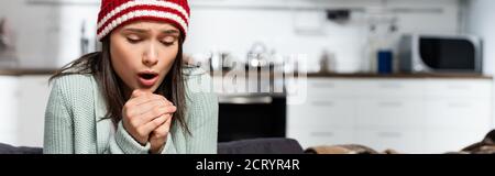 image horizontale de la femme de congélation en bonnet tricoté soufflant dessus les mains se sont trempées dans la cuisine froide Banque D'Images