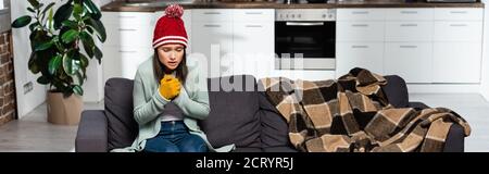 orientation panoramique de la femme de congélation en bonnet et gants tricotés assis sur un canapé près d'une couverture en tissu écossais dans la cuisine froide Banque D'Images