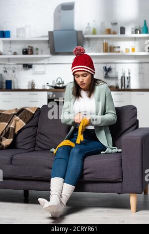 femme de congélation en bonnet tricoté et chaussettes mettant au chaud gants assis sur un canapé dans une cuisine froide Banque D'Images