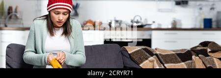 en-tête de site web de la femme de congélation en tricoté chapeau mettant sur gants chauds tout en étant assis sur le canapé dans la cuisine froide Banque D'Images