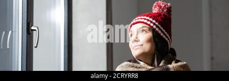 en-tête de site web de la jeune femme dans chaud tricoté chapeau regardant en vous tenant près de la fenêtre à la maison Banque D'Images
