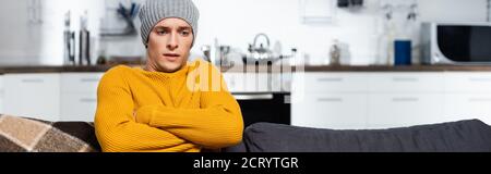 récolte horizontale d'homme froid dans un pull chaud et un chapeau assis avec des bras croisés dans la cuisine froide Banque D'Images