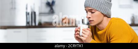 en-tête de site web de jeune homme en bonnet tricoté tenant la tasse de boisson chaude dans la cuisine froide Banque D'Images