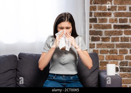 femme malade éternuant dans une serviette en papier tout en étant assise sur un canapé près d'une tasse de boisson chaude Banque D'Images