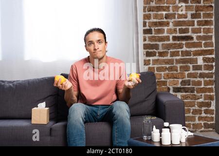 homme malade tenant des citrons frais tout en étant assis sur le canapé près table de chevet avec médicaments Banque D'Images