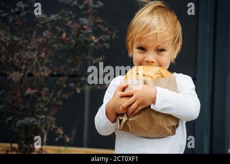 Joyeux garçon aux cheveux blonds tenant du pain croustillant dans les mains, à l'extérieur dans la rue