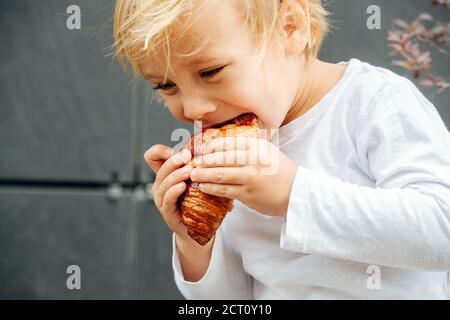 petit garçon glouton aux cheveux blonds en train de manger un croissant croustillant à l'extérieur sur la rue Banque D'Images