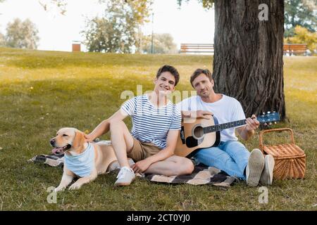 garçon et père adolescents assis sur une couverture près de golden retriever sous le tronc de l'arbre Banque D'Images