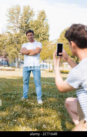 foyer sélectif de l'homme debout avec des bras croisés pendant que l'adolescent garçon prenant des photos et tenant son smartphone dans le parc Banque D'Images