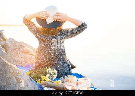 Une belle jeune fille rencontre l'aube lors d'un pique-nique avec un petit déjeuner romantique. La fille est assise face à la mer et au soleil, tenant son chapeau avec ses mains. Banque D'Images