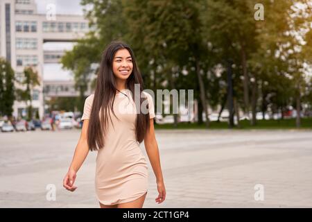 Fille asiatique dans une belle robe beige ajustée marche à travers les rues de la ville et regarde loin. Espace libre pour texte tendance femme Outdoors Sum Banque D'Images