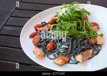 Pâtes spaghetti noires colorées à l'encre de calmar, sauce crème, saumon, tomates et salade de roquette dans une assiette blanche sur une table de jardin rustique sombre, se Banque D'Images