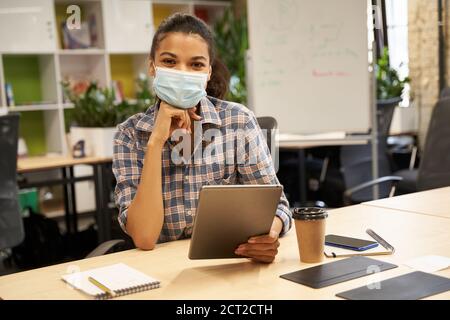 Jeune femme, employée de bureau portant un masque de protection, tenant une tablette numérique et regardant l'appareil photo tout en travaillant dans un bureau moderne. Crise de 19, crise de l'entreprise et de la covid, prise de distance sociale Banque D'Images