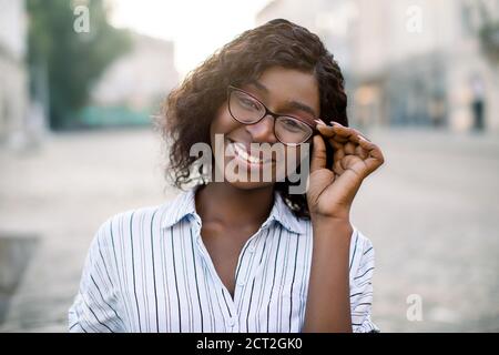 Portrait urbain en gros plan d'une femme africaine positive élégante en chemise rayée décontractée, touchant ses lunettes, souriant à l'appareil photo marchant à l'extérieur Banque D'Images