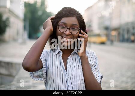 Gros plan portrait d'une femme africaine souriante en lunettes, marchant dans la vieille ville avec un téléphone portable, parlant avec son ami et souriant à l'appareil photo. Personnes