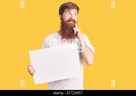 Beau jeune homme à tête rouge avec une longue barbe tenant une bannière vide vierge sérieux visage pensant à la question avec la main sur le menton, réfléchi à la confus Banque D'Images