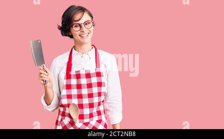 Belle jeune femme aux cheveux courts portant un tablier de boulanger professionnel tenir le couteau en position positive, se tenir debout et sourire un confiant Banque D'Images