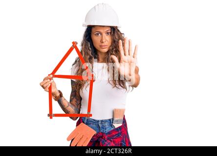 Jeune femme hispanique avec tatouage portant architecte hardhat holding projet de construction avec main ouverte faisant signe d'arrêt avec une expression sérieuse et confiante, Banque D'Images