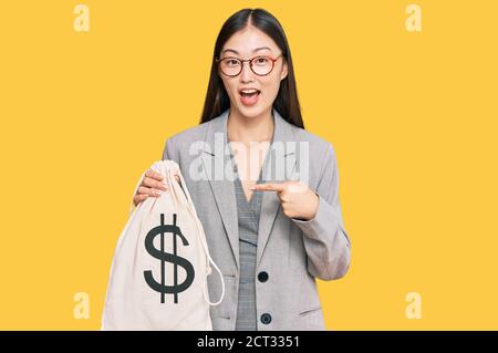 Jeune femme chinoise portant un costume d'affaires tenant un sac de dollars souriant contenchez-vous de pointer avec votre main et votre doigt Banque D'Images