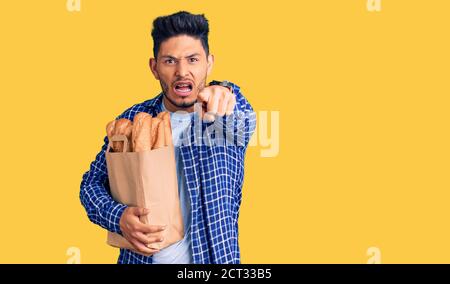 Beau jeune homme latino-américain tenant un sac de papier avec du pain pointant mécontent et frustré à l'appareil photo, furieux et furieux avec vous Banque D'Images
