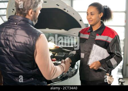 Jeune ouvrier de service de voiture avec le document technique secouant la main de client masculin Banque D'Images