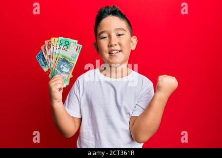 Petit garçon hispanique enfant tenant des dollars australiens criant fier, célébrant la victoire et le succès très excité avec le bras levé Banque D'Images