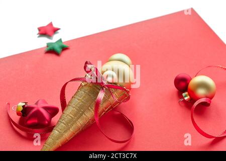 Cornet de glace gaufré doré avec boules dorées de Noël, étoiles et rubans rouges. Arrangement de Noël sur papier rose isolé sur blanc. Banque D'Images