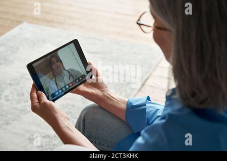 Vue sur l'épaule d'une vieille femme qui appelle un médecin virtuel sur une tablette à la maison. Banque D'Images