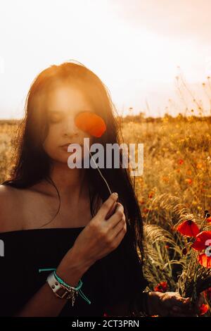 Portrait d'été de belle femme ukrainienne bronzée, brunette avec de longs cheveux foncés fermant un oeil avec des fleurs de pavot rouge et un bouquet à la main dans les s Banque D'Images