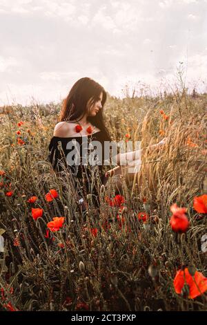 Harmonie de la nature. Belle jeune femme brunette avec des cheveux longs brun foncé dans les épaules robe noire assis dans le profil dans un coquelicots sauvages fleurs et Banque D'Images