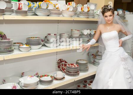 mariée debout à côté des étagères avec des plats et des assiettes dedans section ustensiles de cuisine d'un magasin Banque D'Images