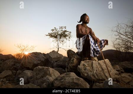 Un homme Himba est assis sur un rocher pendant que le soleil se couche à la fin de la journée à Epupa, dans la région de Kunene, en Namibie. Banque D'Images