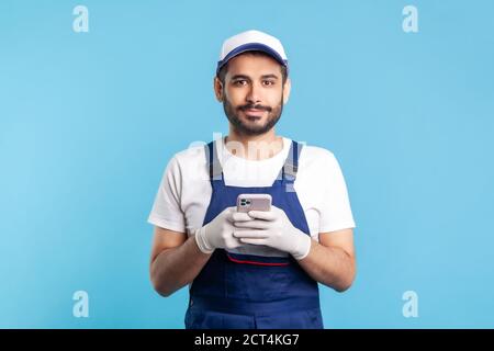 Handyman dans des combinaisons et des gants tenant le téléphone mobile, en utilisant le messager de téléphone cellulaire pour accepter la commande en ligne comme services de livraison, de réparation et d'entretien. i Banque D'Images