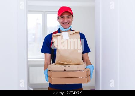 Livreur employé dans le t-shirt rouge casquette uniforme masque gants donner des boîtes à pizza de commande d'aliments isolé sur fond jaune studio. Mise en quarantaine du service Banque D'Images