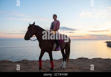 Une fille blonde est assise sur un cheval contre le fond de la mer et un coucher de soleil lumineux. Banque D'Images