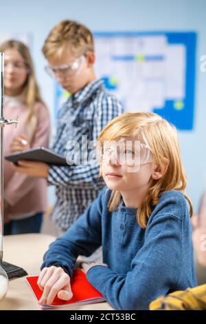 Portrait d'un petit garçon mignon portant des lunettes de sécurité, assis à son bureau d'école dans la salle de classe des sciences de chimie, Smiles heureusement. Portrait en gros plan Banque D'Images