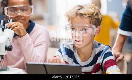 École élémentaire Science salle de classe: Garçon utilise l'ordinateur numérique tablette pour vérifier l'information sur l'Internet tandis que l'enthousiaste mignon petite fille utilise Banque D'Images