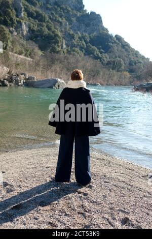 vue arrière de la femme avec de courts cheveux orange et habillée Dans une tenue d'hiver élégante se dresse sur la banque de galets de la rivière Herault vue sur la rivière qui coule Banque D'Images
