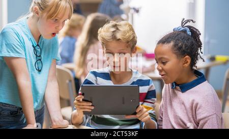 Cours de sciences informatiques de l'école élémentaire : deux filles et garçons utilisent un ordinateur de tablette numérique avec logiciel de réalité augmentée, ils sont enthousiastes, plein de Banque D'Images