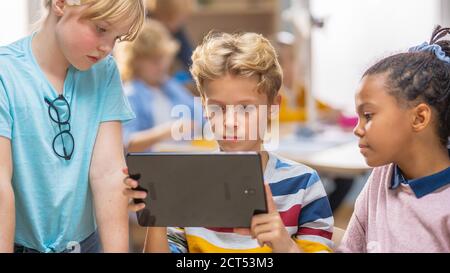 Cours de sciences informatiques de l'école élémentaire : deux filles et garçons utilisent un ordinateur de tablette numérique avec logiciel de réalité augmentée, ils sont enthousiastes, plein de Banque D'Images