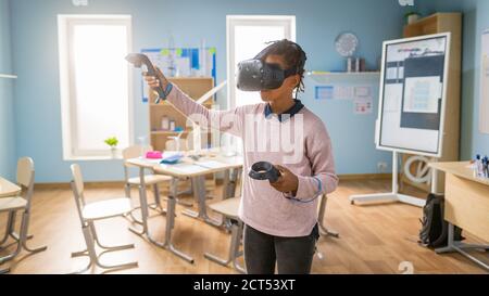 Cours de sciences informatiques à l'école élémentaire : une fille adorable porte un casque de réalité augmentée et utilise des contrôleurs apprend des leçons dans la réalité virtuelle excitée Banque D'Images