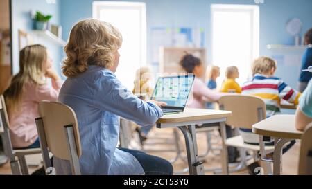 Cours de sciences de l'école élémentaire : petit garçon utilise un ordinateur portable avec écran montrant le logiciel de programmation. Un professeur de physique explique la leçon à une classe diversifiée Banque D'Images