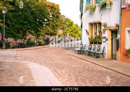 Vue sur la rue pavée française romantique avec café, lampes de rue et parc en arrière-plan. Tables et chaises vides à l'extérieur du restaurant, Colmar Banque D'Images